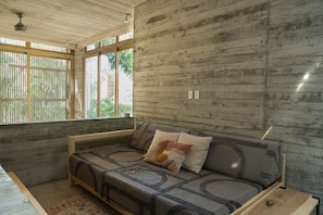 Una habitación con techo de madera, pared de concreto, un sofá con cojines y una ventana con vista a follaje.