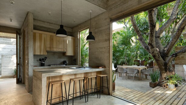 Una cocina moderna con una isla grande, taburetes de bar, lámparas colgantes y vistas a un patio exterior con zona de comedor y un árbol.