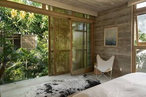 Quarto com uma grande janela, uma cadeira, uma cama e vista para vegetação exuberante.