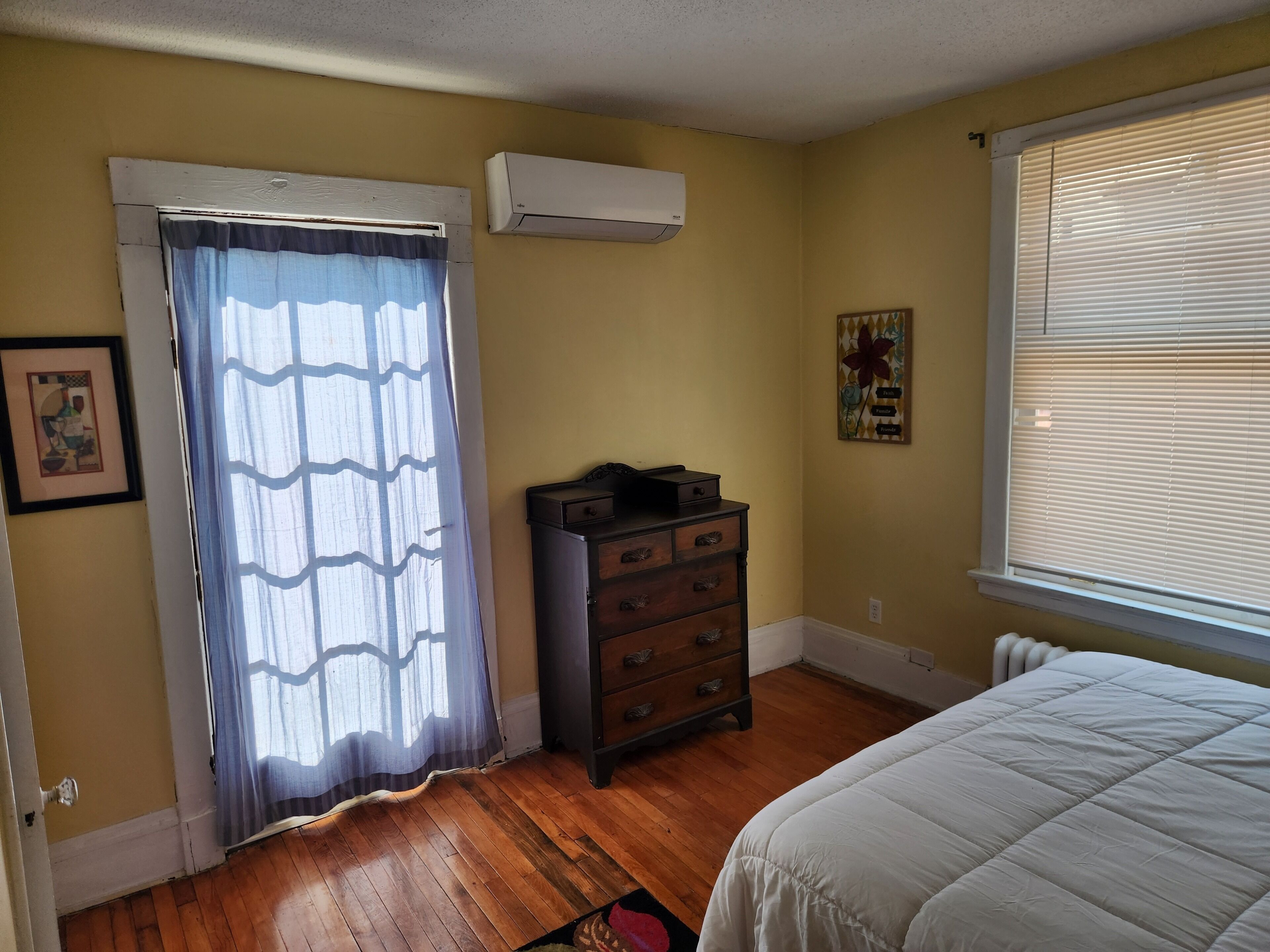Un dormitorio con suelo de madera, cama, cómoda, unidad de aire acondicionado, ventana con persianas y puerta con cortina.