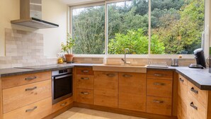 Una cocina con armarios de madera, una campana extractora de acero inoxidable, un horno empotrado, un ventanal con vistas a árboles y una encimera con cafetera.