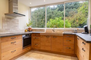 A kitchen with wooden cabinets, a stainless steel range hood, a built-in oven, a large window with a view of trees, and a countertop with a coffee maker. - EcoGîte Cévénola (sleeps 3) (MOLEZON)