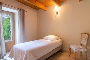 A bedroom with a bed, a chair, a window with curtains, and a wall-mounted light fixture. - EcoGîte Cévénola (sleeps 3) (MOLEZON)
