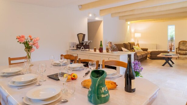 Un comedor con una mesa dispuesta para cuatro, una cocina al fondo y una sala de estar con un sofá y una lámpara.