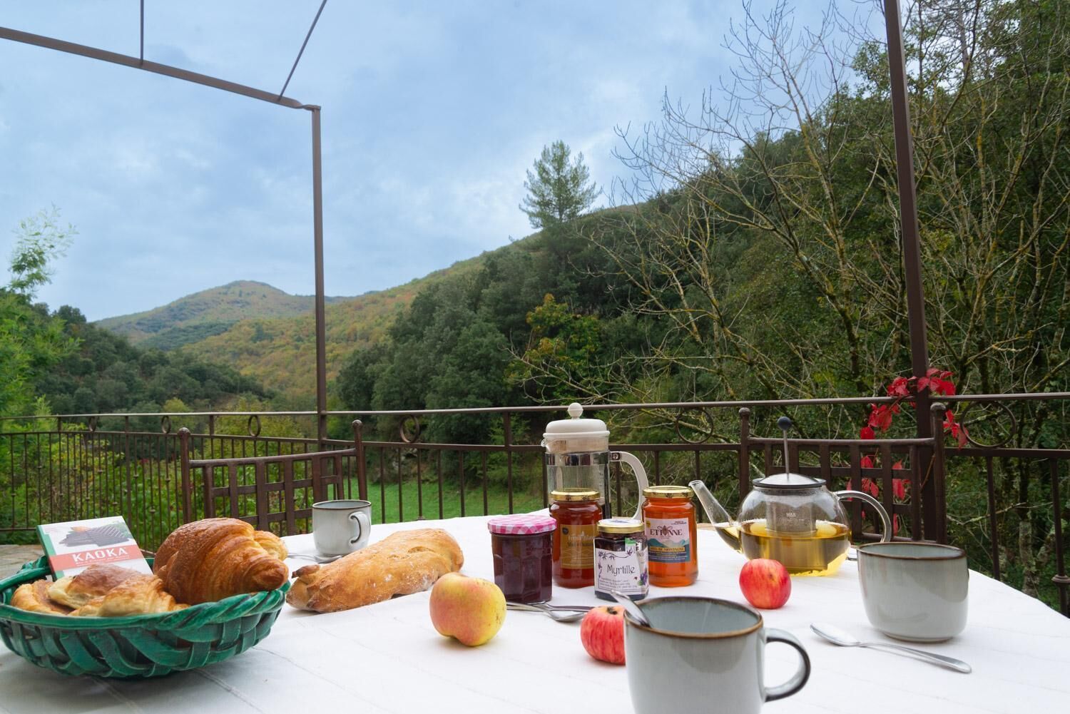 Una mesa con comida y bebidas, una cesta de pan y vistas a una ladera boscosa.