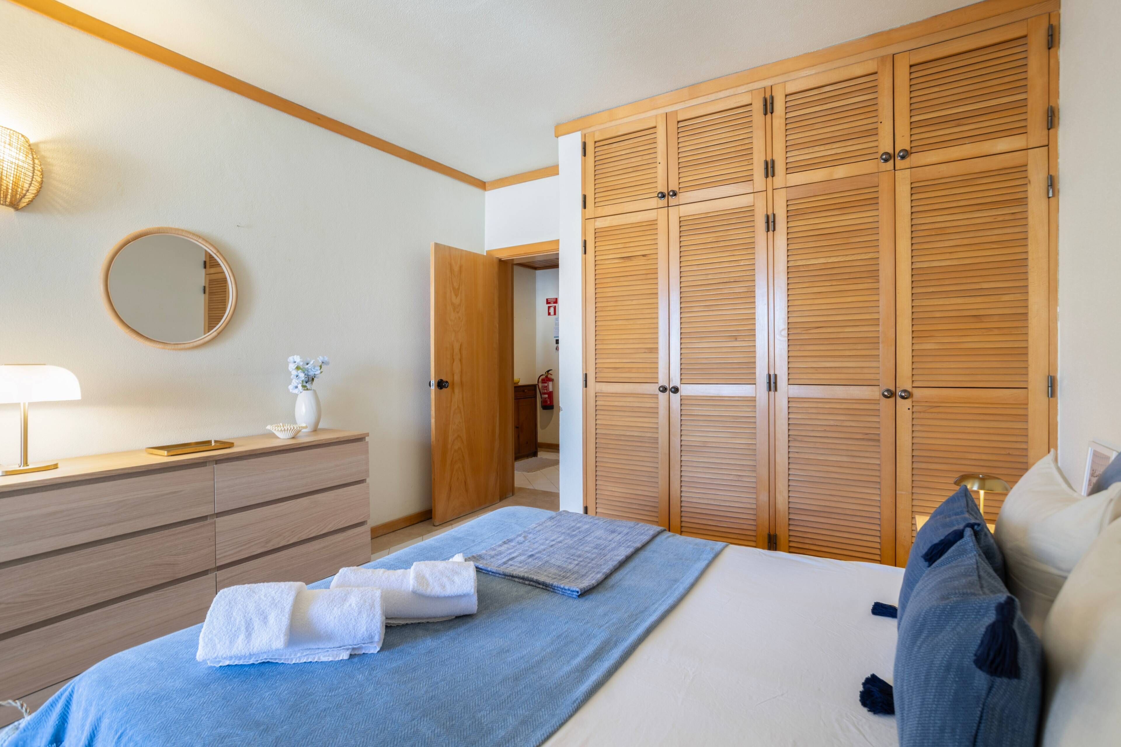 Ein Schlafzimmer mit einem Bett, einem Nachttisch mit Lampe, einer Kommode, einem Spiegel und einem Holzschrank.