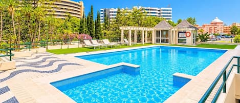 A swimming pool with lounge chairs and a gazebo, surrounded by greenery and buildings.