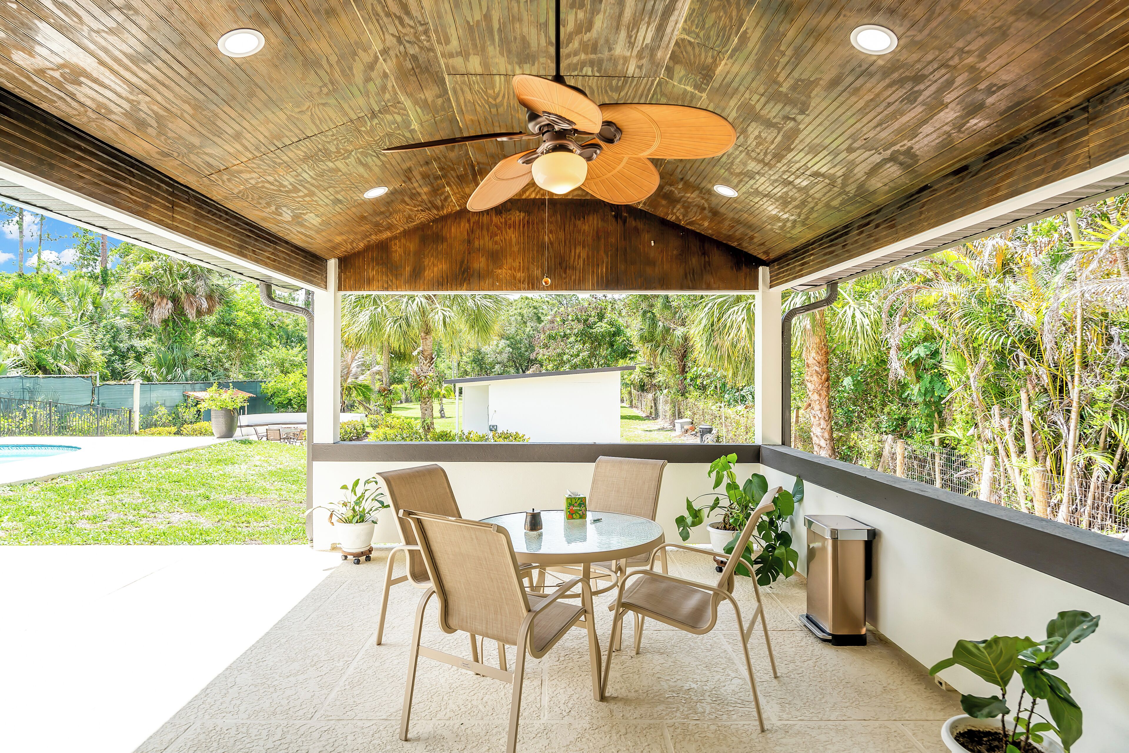 A covered patio with a ceiling fan, a round table, and chairs, overlooking a lush garden.