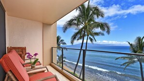 A balcony with wicker chairs, a small table with a flower vase, and a view of the ocean and palm trees. - ~MAHANA 505~Ocean Front Condo with A/C & Beautiful Island Views in Kaanapali (Lahaina)