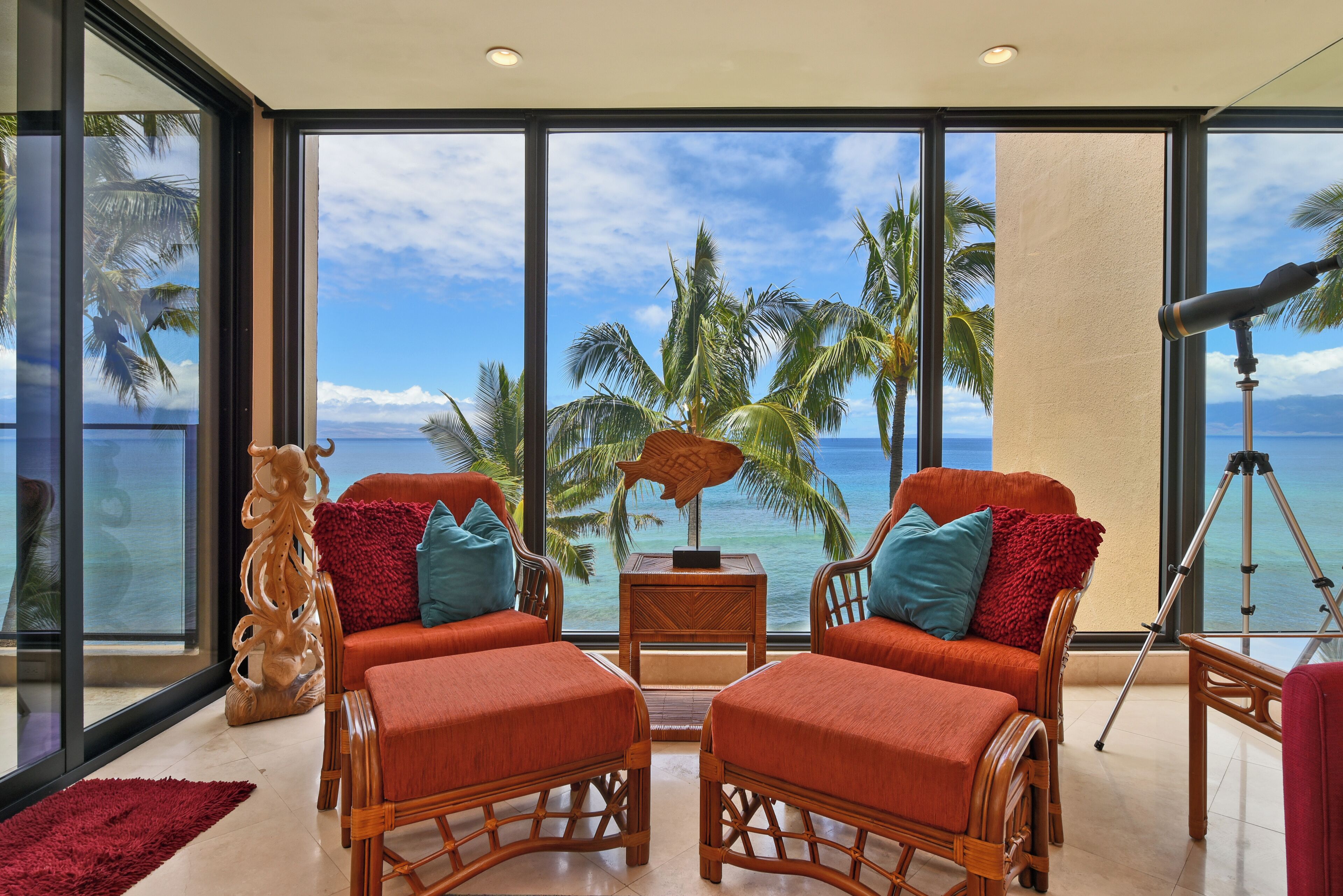 A room with a large window overlooking the ocean, featuring wicker furniture and a red rug.