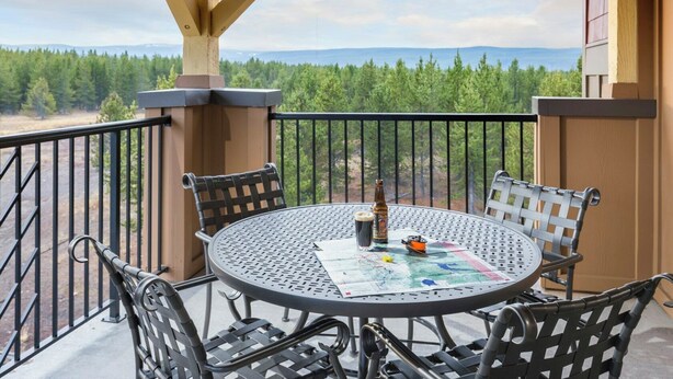 Ein runder Metalltisch mit vier Stühlen, einer Bierflasche und einer Karte auf einem Balkon mit Blick auf Bäume und Berge.