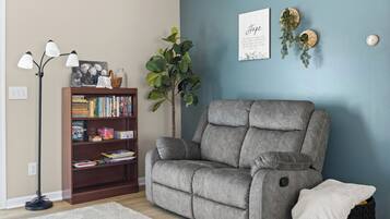 A living room with a grey sofa, a wooden bookshelf, a floor lamp, a large plant, and two wall art pieces.
