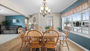 A dining room with a wooden table and chairs, a chandelier, and a view of the outside through large windows.