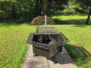A metal outdoor grill with a tree and animal design, set on a stone base in a grassy area with trees and a stack of firewood in the background.