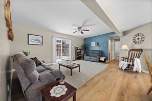 A spacious living room with a sofa, a coffee table, and a rocking chair. There is a bookshelf and a large window with a view outside. - Private Backyard & Deck · Family-Friendly (Penn Yan)