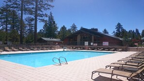 En swimmingpool med solstolar, en byggnad med ett rött tak och höga träd i bakgrunden.