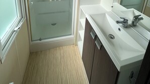 A bathroom with a glass shower enclosure, a white sink with a mirror, and wooden cabinets. - Base de Loisirs du Colvert - Mobile Home Évasion (Bonzée)