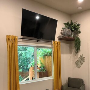 Ein Wohnzimmer mit an der Wand montiertem Fernseher, einem Fenster mit gelben Vorhängen, einem grünen Sessel und einem Regal mit Pflanzen.