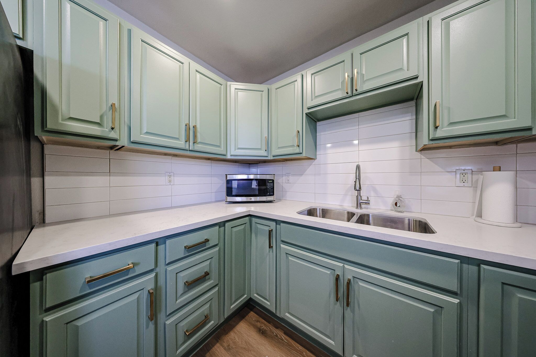 A kitchen with teal cabinets, white countertops, a stainless steel sink, and a microwave.
