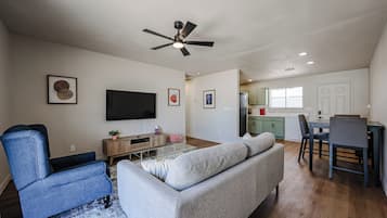 A living room with a sofa, a TV, a ceiling fan, and a dining area with chairs and a table.