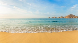 A beach with clear water, sandy shore, and rocky formations in the distance. - Cabo Beachfront Escape with Spacious 2BR Suite & Resort Pools (Cabo San Lucas)