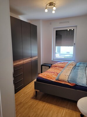Ein Schlafzimmer mit einem Bett, einem Kleiderschrank, einem Nachttisch, einem Fenster mit Jalousien und einer Deckenleuchte.