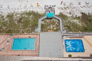 Luftaufnahme eines Resorts mit zwei Schwimmbecken, einem Strandbereich und einem überdachten Gang.