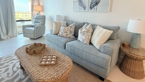 Uma sala de estar com um sofá cinzento, um banquinho de vime, uma poltrona cinzenta e vista da cidade através de uma grande janela.