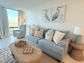 Ein Wohnzimmer mit grauem Sofa, einem Korbsessel, einem grauen Sessel und Blick auf die Stadt durch ein großes Fenster.