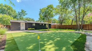 A residential backyard with artificial grass, a golf hole, and a house with a garage. - Modern Dallas Retreat • Golf • Speakeasy• Fire Pit (Dallas)