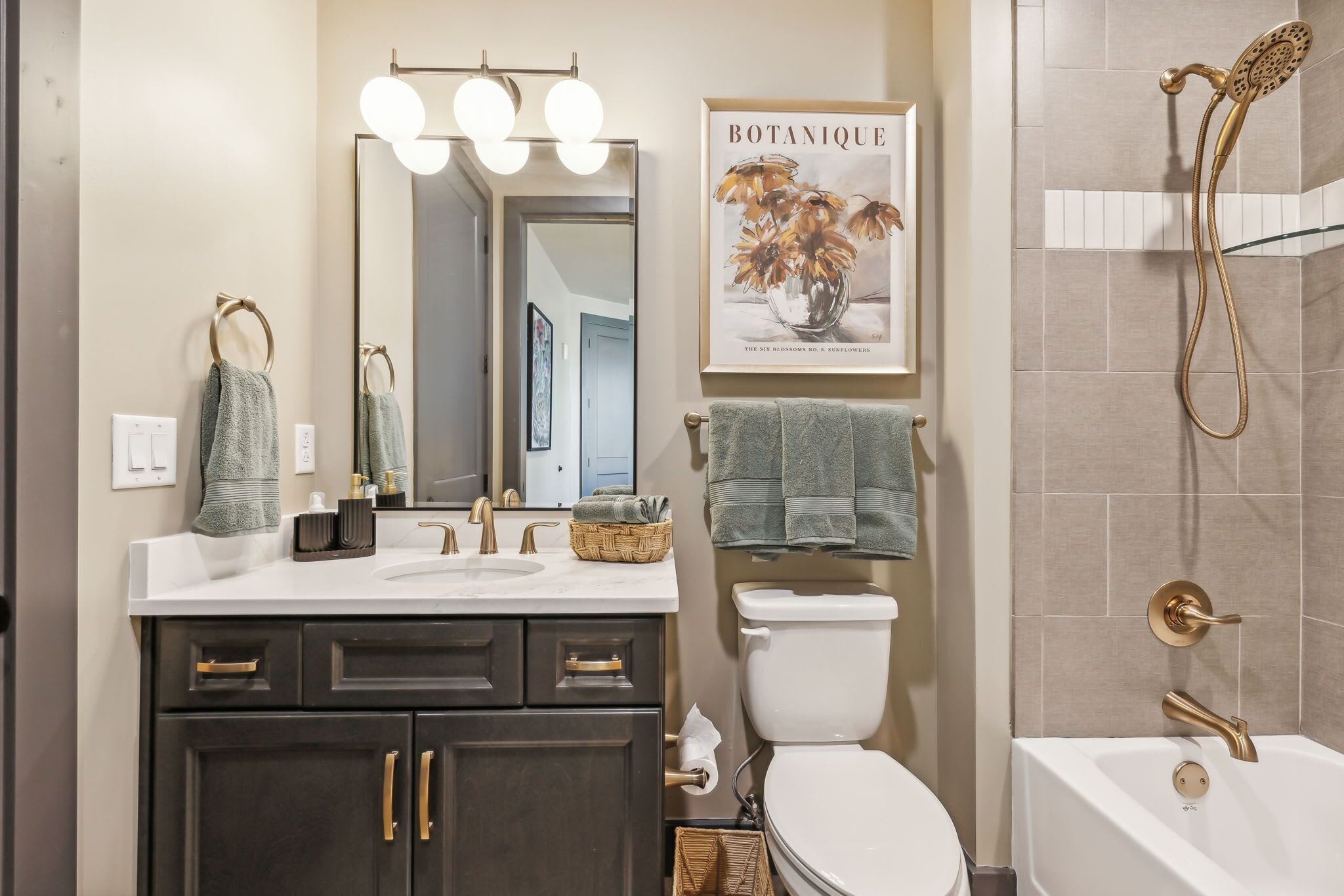 Une salle de bain avec un lavabo blanc, des meubles sombres, des toilettes, une douche dotée d’un pommeau de douche doré et une estampe botanique encadrée au mur.