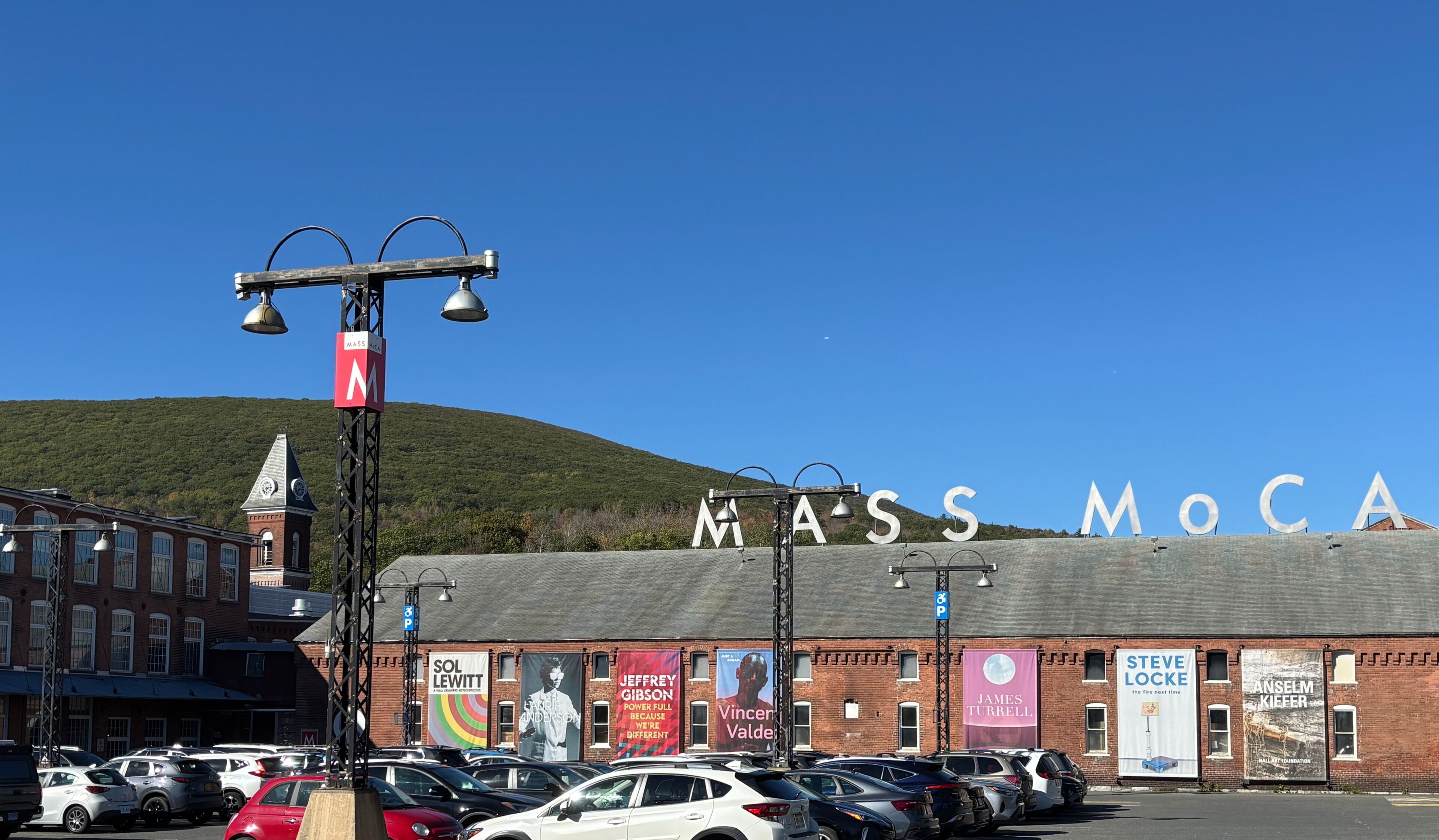 A parking lot with cars, a building with 'MASS MoCA' signage, and banners advertising various artists and events.