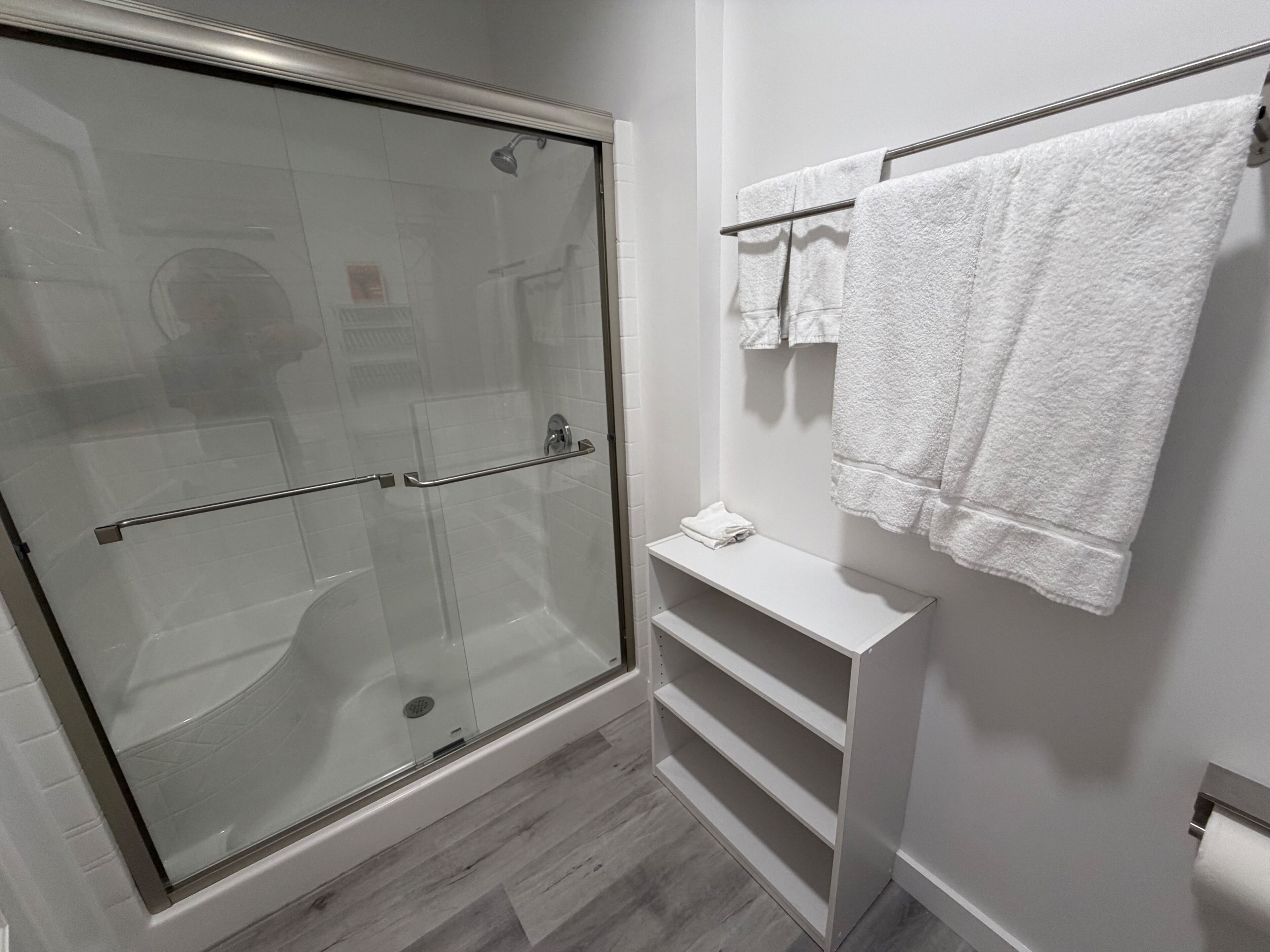 Un baño moderno con cabina de ducha de vidrio, paredes con azulejos blancos, un perchero blanco y un estante blanco.