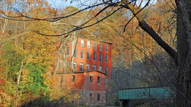 A red brick building with multiple windows, surrounded by autumn foliage and a river.