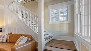 Una escalera con balaustres blancos, un sofá de cuero marrón con cojines estampados y una ventana con contraventanas.