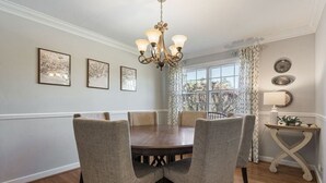 Ein Esszimmer mit einem runden Tisch, sechs Stühlen, einer Deckenleuchte, gerahmten Bildern an der Wand und einem fenster mit Vorhängen.