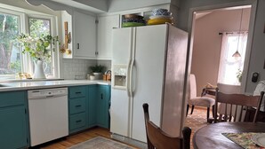 Une cuisine avec des meubles blancs, un plan de travail de couleur turquoise, un réfrigérateur blanc et un coin repas avec une table et des chaises en bois.
