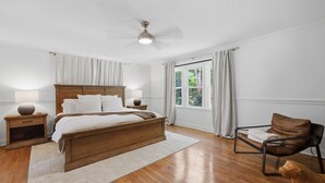 Quarto com cama de madeira, mesa de cabeceira, uma cadeira, um cão e janela com cortinas.