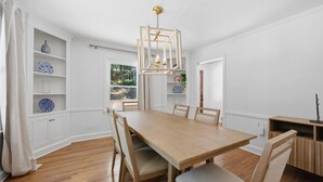 Uma sala de jantar com uma mesa de madeira, cadeiras, uma luminária pendente e paredes brancas.