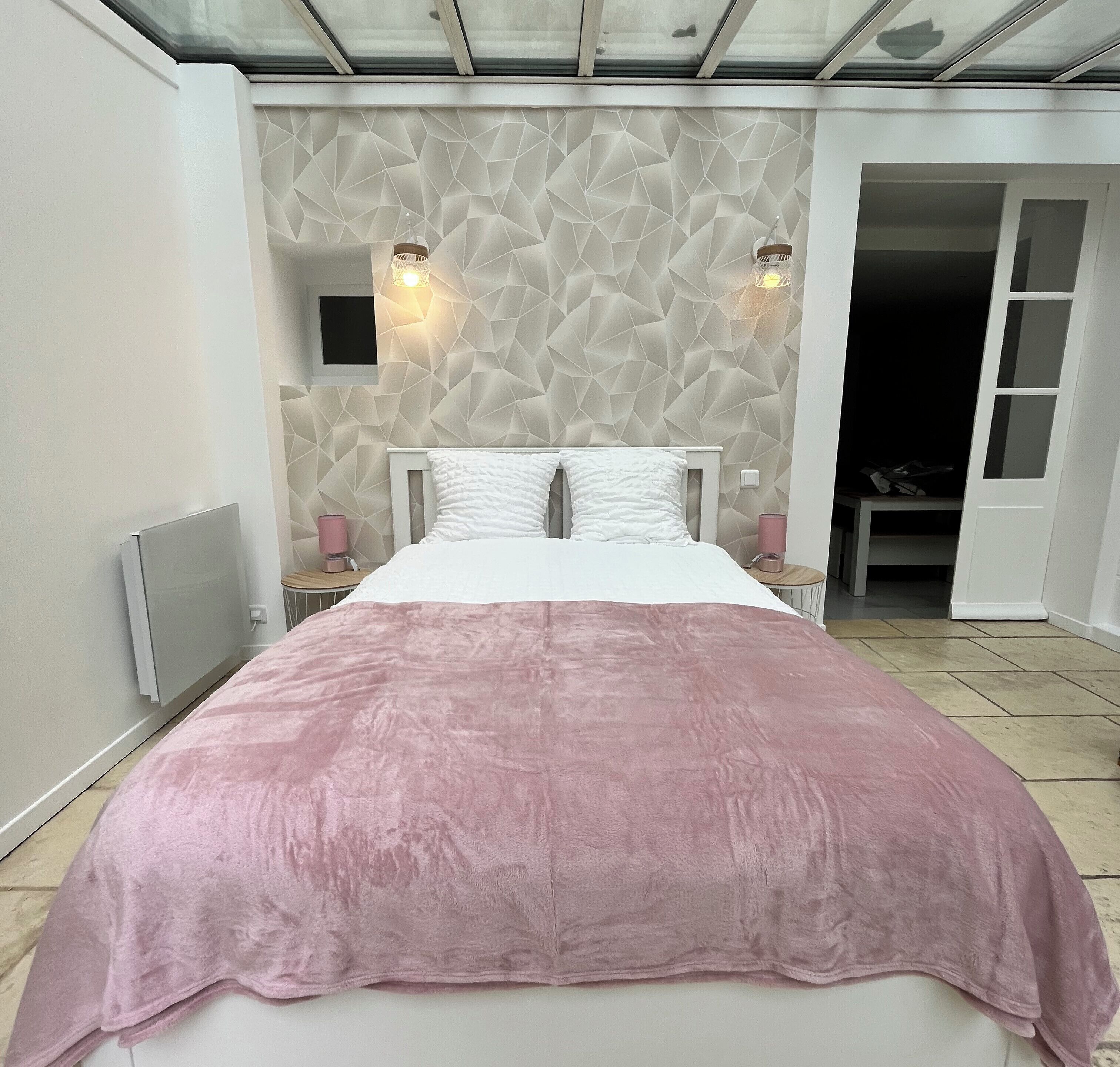 A bedroom with a bed, a geometric-patterned wall, two wall-mounted lights, a pink blanket, and a door leading to a closet.