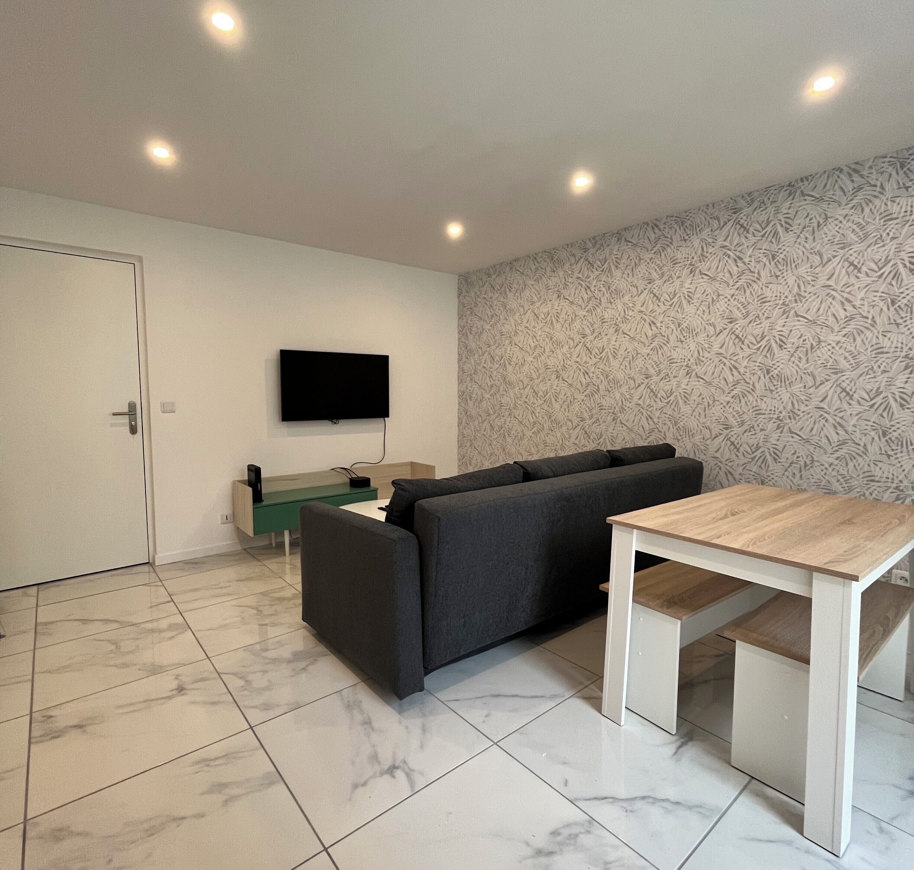 Ein modernes Wohnzimmer mit einer Couch, einem an der Wand befestigten Fernseher, einem kleinen Tisch und einer Wandtapete mit Muster.