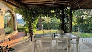 Un patio exterior con mesa y sillas, rodeado de follaje y vistas al campo.
