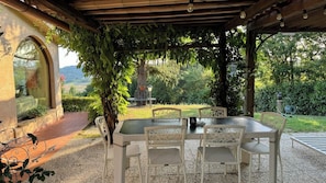 Um pátio externo com mesa e cadeiras, cercado por vegetação e vista para o campo.