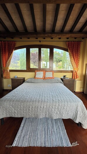 Quarto com cama grande, vigas de madeira no teto, janela com cortinas e mesa de cabeceira com abajur.