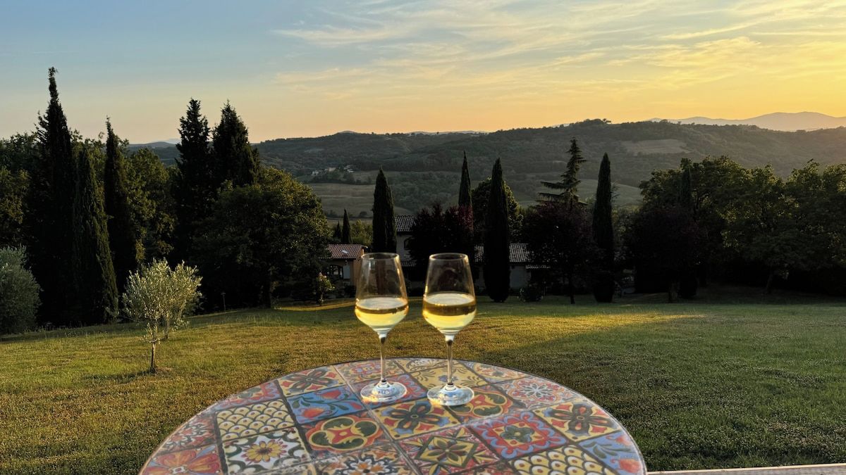 Una mesa con dos copas de vino sobre un mantel con un estampado colorido, con un paisaje de árboles y colinas al fondo.