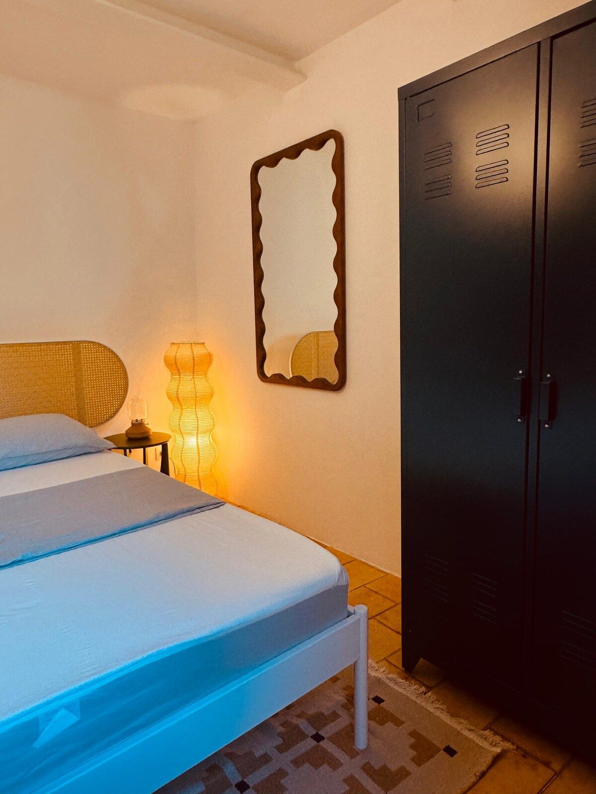 Un dormitorio con una cama, un armario, un espejo adosado a la pared y una lámpara de noche.