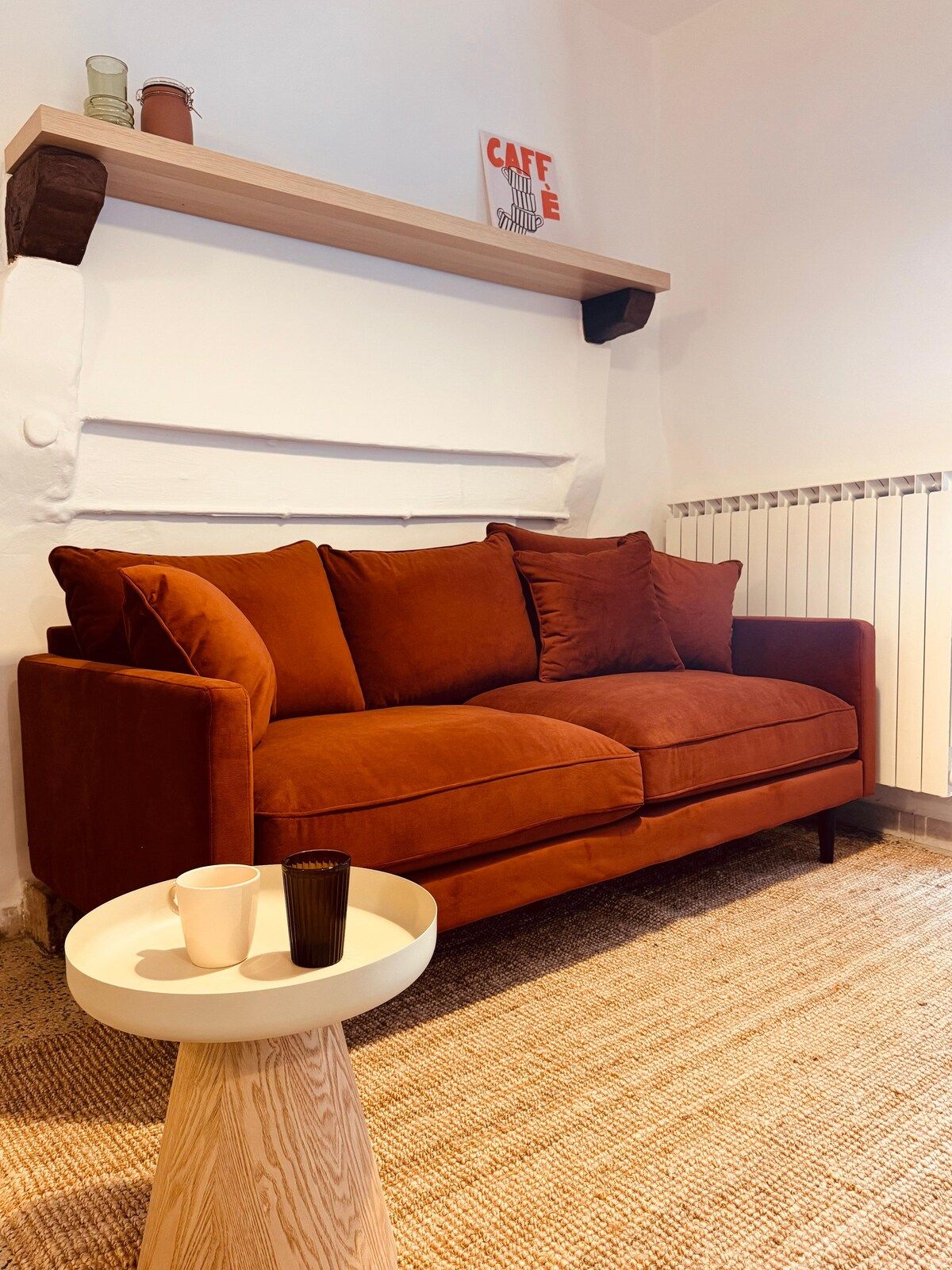 A room with a brown sofa, a small round table with a cup and a vase, a shelf with jars, and a radiator.