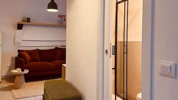 Un espacio habitable compacto con sofá, reposapiés e iluminación colgante. Una puerta da acceso a un baño con ducha e inodoro.