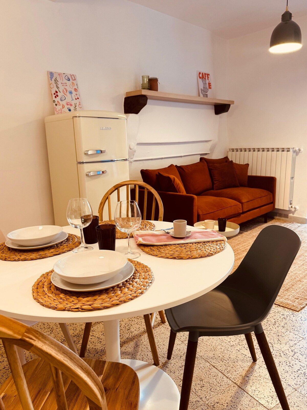 Un coin repas avec une table ronde dressée pour deux personnes, un réfrigérateur blanc, un canapé marron et un luminaire suspendu.