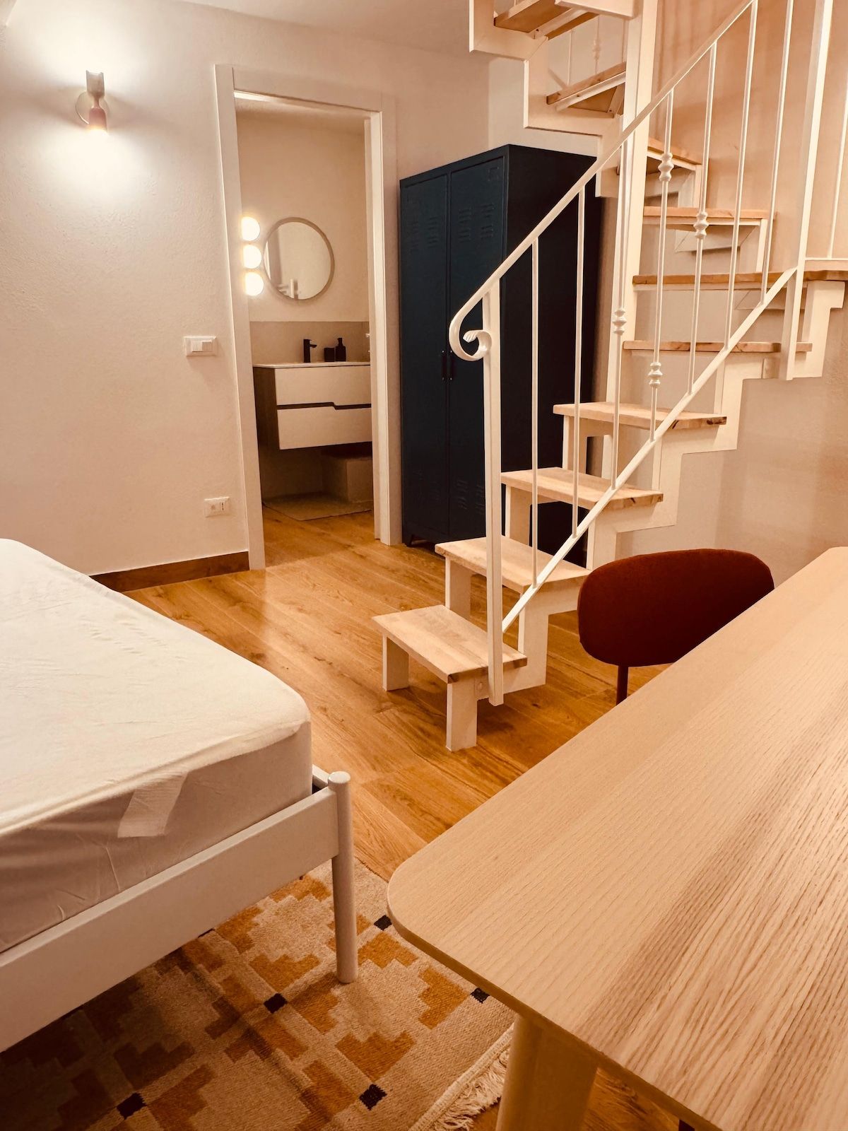 Een slaapkamer met een bed, bureau, stoel, een trap en een badkamer zichtbaar door een geopende deur.